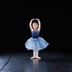 A young dancer in a navy leotard and light blue tutu standing in first position with arms raised overhead in a circle, on a stage with a black backdrop.