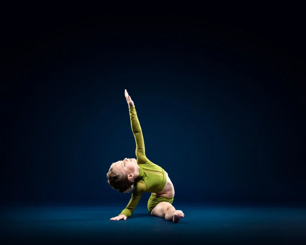 A teenage dancer in a light blue gi-style top and pants balanced on one foot with the other leg extended high in a controlled side kick, against a black studio backdrop