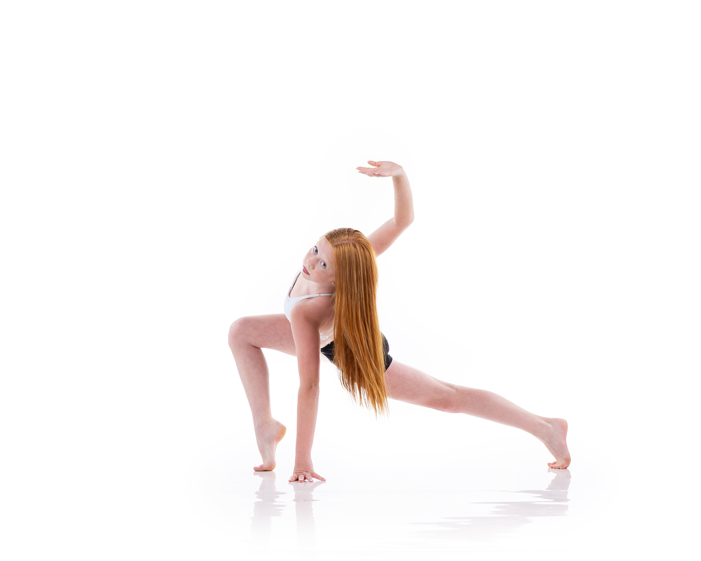 A young dancer with long red hair in a white top and black shorts in a deep lunge, one arm curved overhead, against a white backdrop.