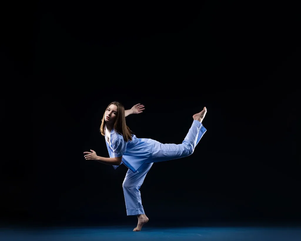 A young dancer in a blue ruffled outfit balanced in a side plank with one leg extended high overhead, against a deep navy studio backdrop