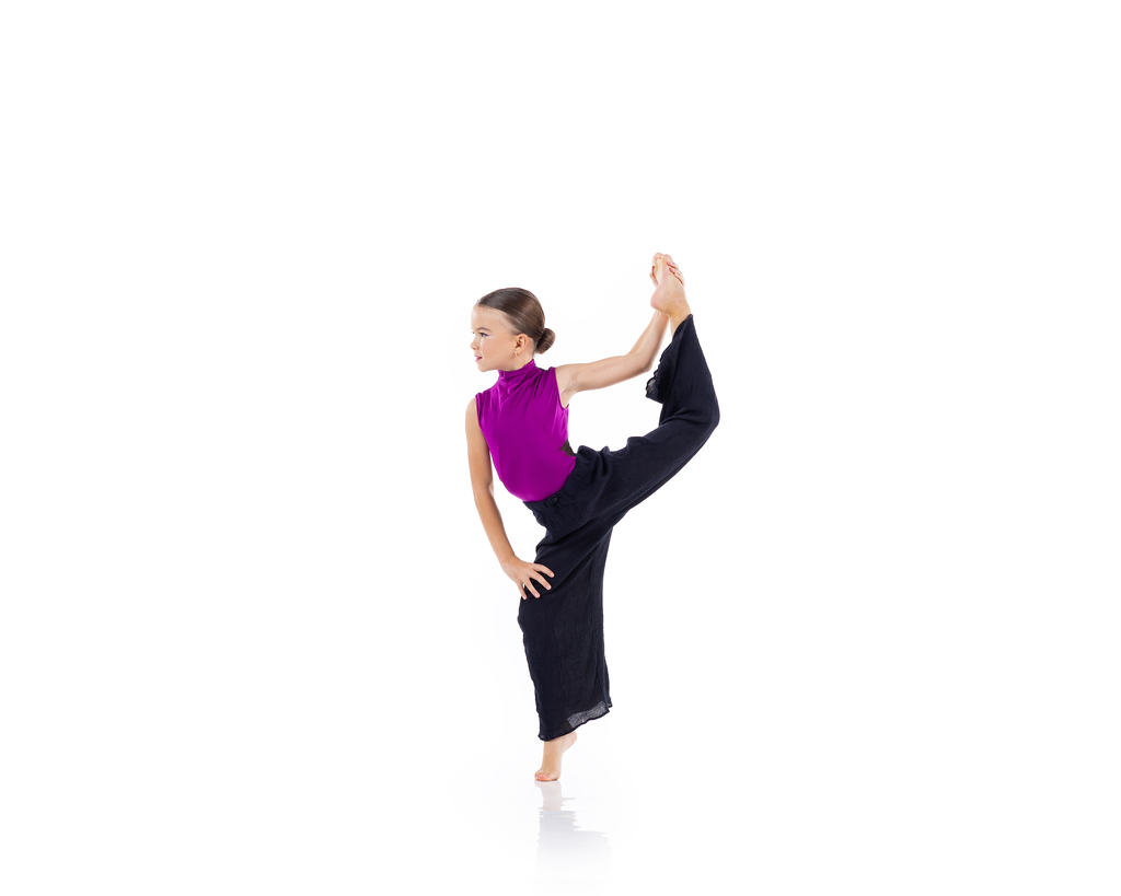 A young dancer in a magenta top and black pants in a standing side stretch, holding her raised foot overhead, against a white backdrop.