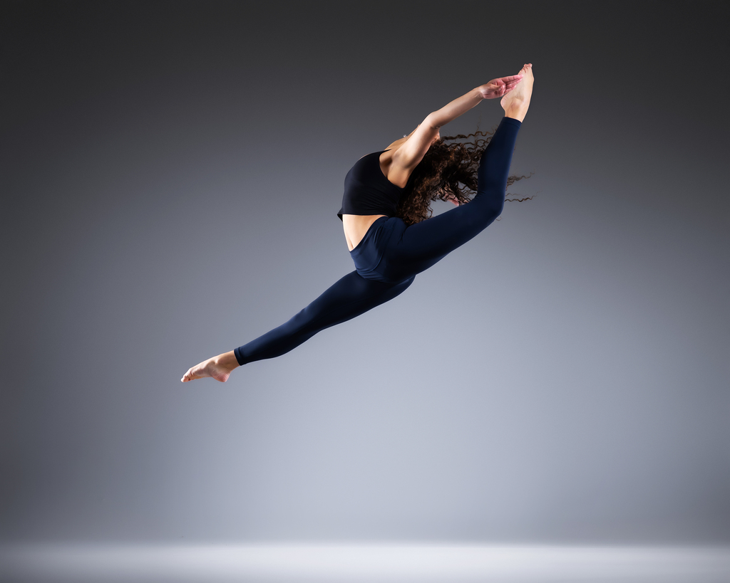 A dancer in a black top and navy leggings captured mid-leap, arching backward with arms and one leg reaching overhead, against a gray backdrop.