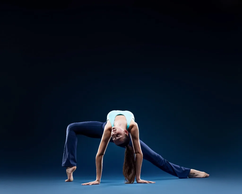 A young dancer in a floral leotard and teal shorts seated on the studio floor reaching back to hold one foot above her head, gaze lifted upward, against a deep navy backdrop